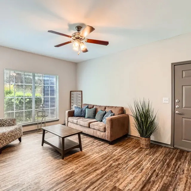 A spacious living room featuring a brown sofa with teal cushions, a light-colored armchair, a coffee table, and a decorative plant. There's a window allowing natural light and a dark door on one side.