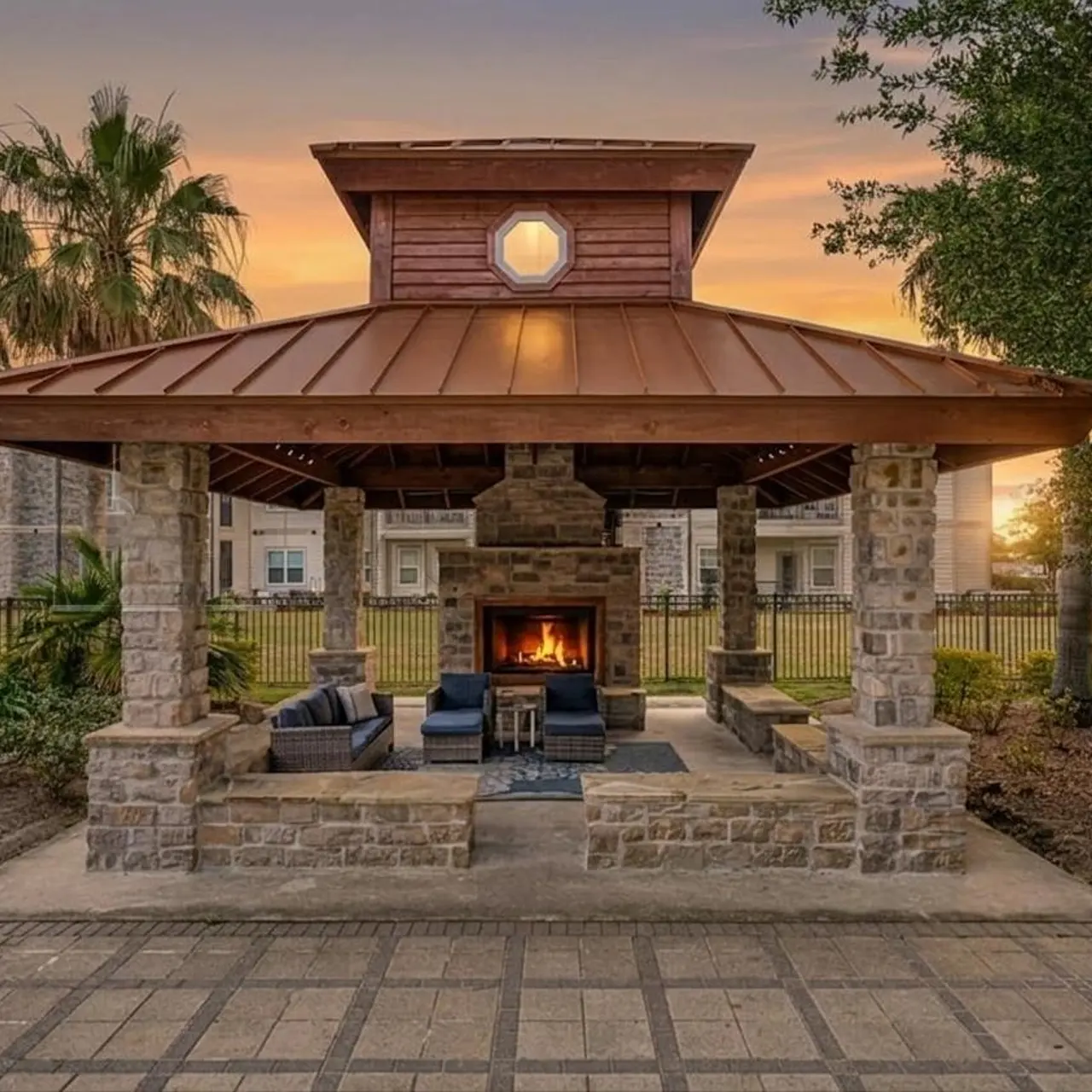 An outdoor pavilion with a stone fireplace, surrounded by lush greenery and palm trees, under a sunset sky.