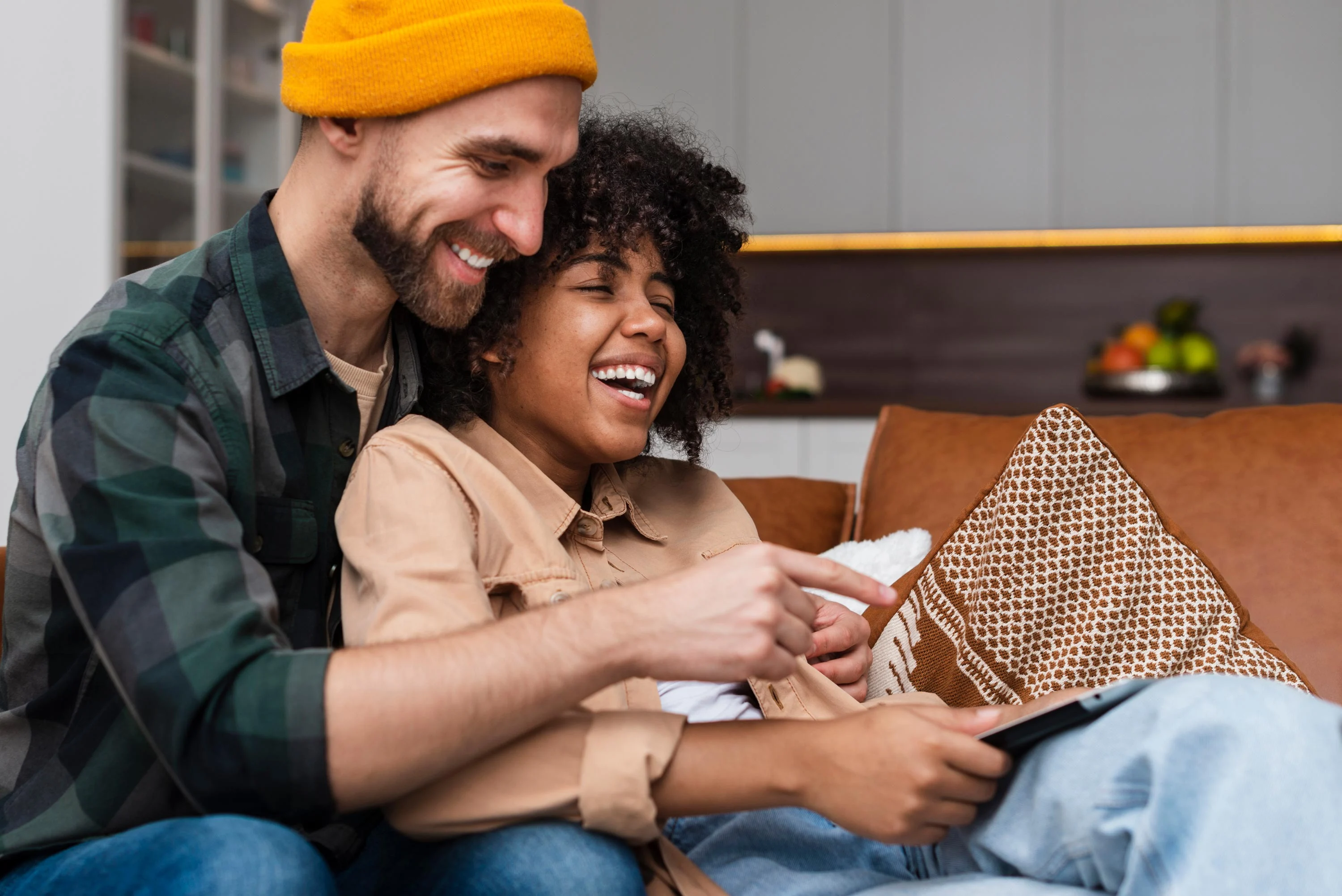 Happy Couple Enjoying Time Together A smiling couple sitting on a couch, with one person pointing at a tablet or phone while the other laughs joyfully.