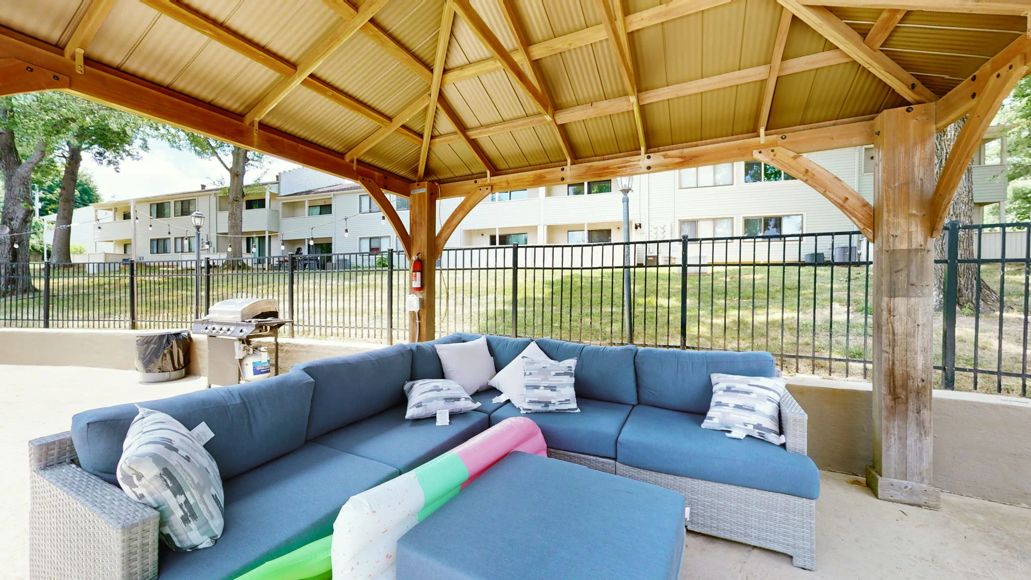 A spacious outdoor lounge area with a covered gazebo, featuring a large sectional sofa, colorful pool float, and a grill in the background.