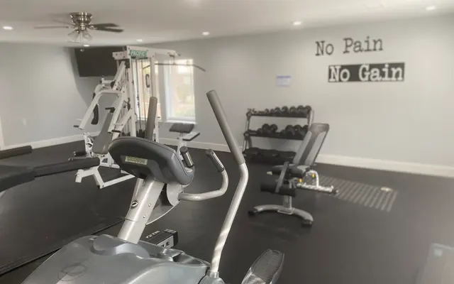 Interior of a home gym featuring various exercise equipment including an elliptical machine, weight bench, and dumbbells. A motivational quote 'No Pain No Gain' is on the wall.