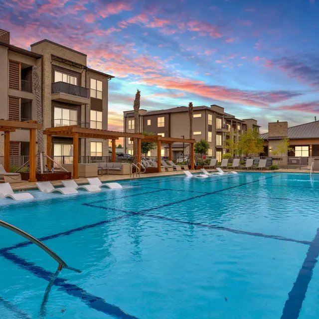 An inviting pool area surrounded by modern apartment buildings, featuring lounge chairs and decorative wooden structures, under a colorful sky at sunset.