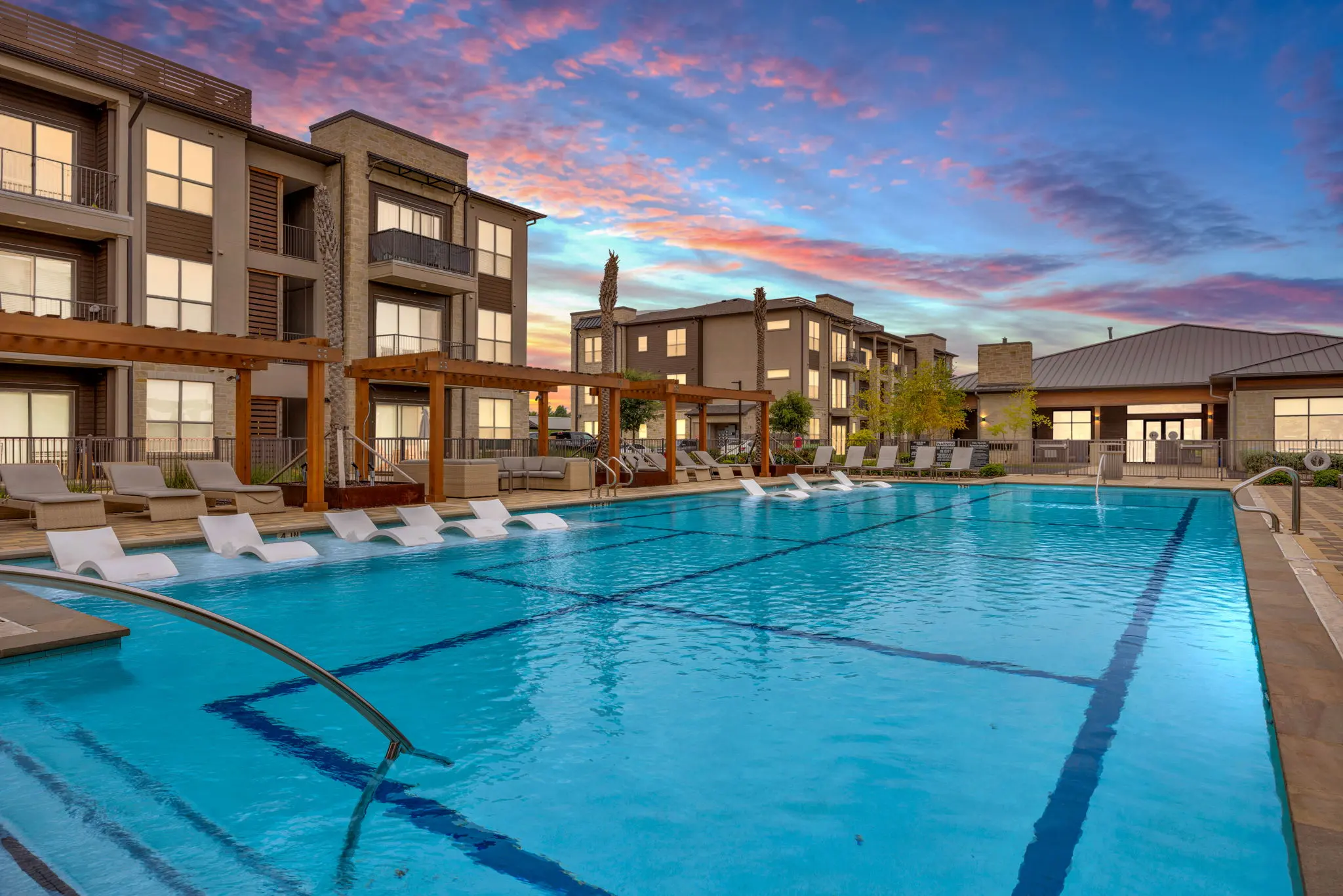 An inviting pool area surrounded by modern apartment buildings, featuring lounge chairs and decorative wooden structures, under a colorful sky at sunset.