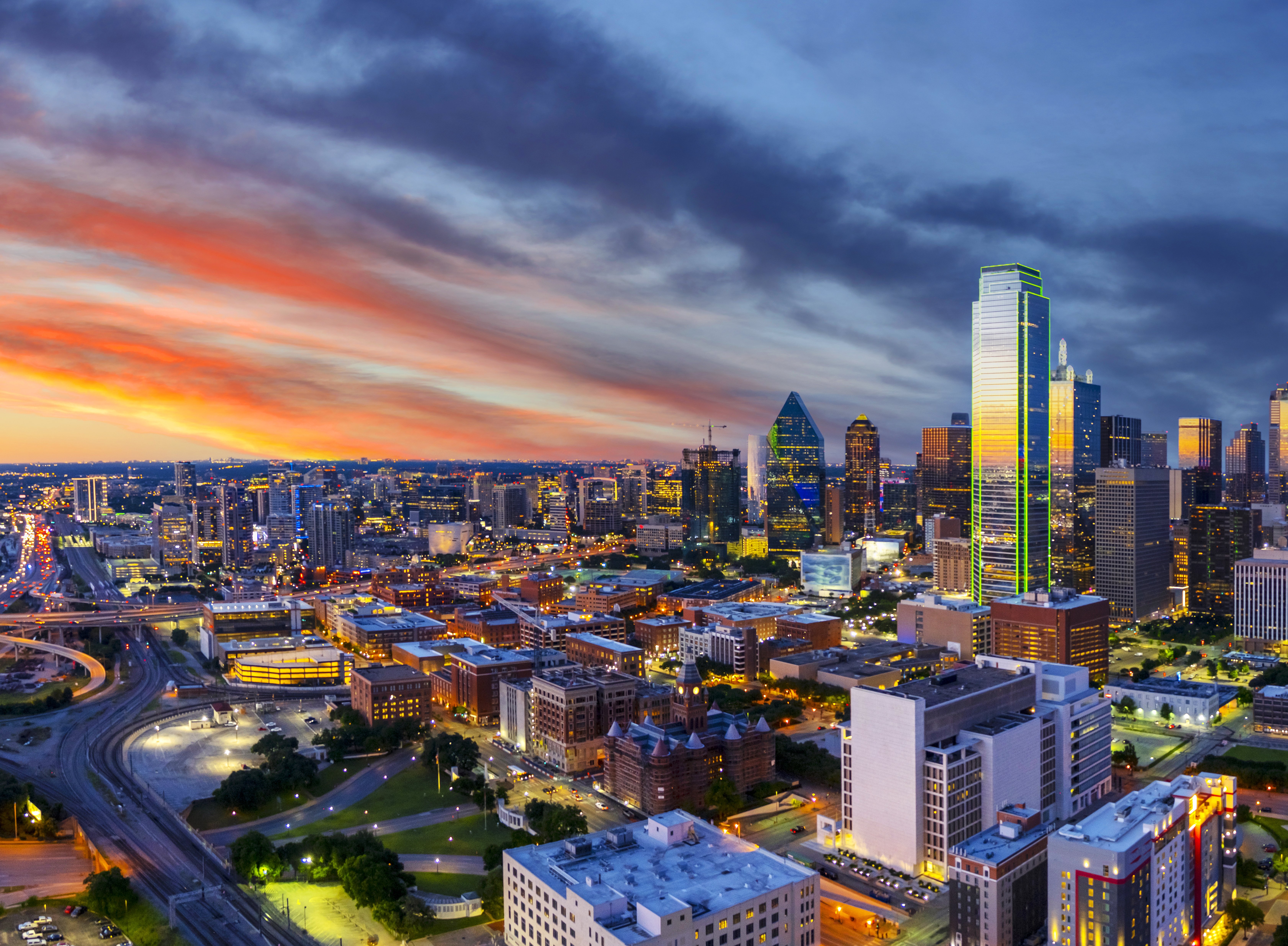 A vibrant sunset view of the Dallas skyline with colorful clouds and city lights reflecting on buildings.