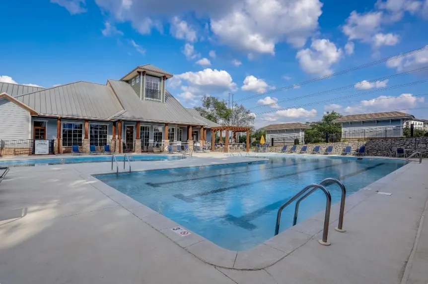 A spacious outdoor swimming pool area with lounge chairs and a community building in the background under a blue sky with clouds.