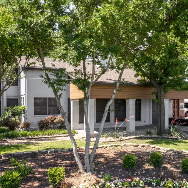 A landscaped area featuring a building partially covered by trees, with flowerbeds in front and a clear sky.
