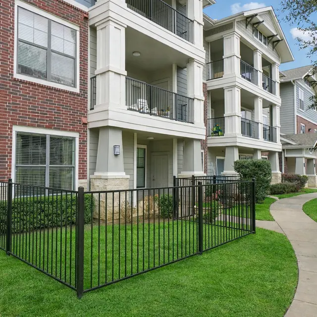 An exterior view of a multi-story apartment complex featuring brick and siding architecture, with a fenced grassy area in the foreground and a curved pathway leading into the background.