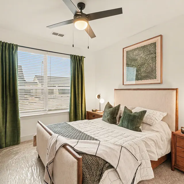 A modern bedroom featuring a comfortable bed with decorative pillows, a wooden headboard, and contemporary lighting. The walls are painted white and large windows are dressed with green curtains, allowing light to fill the space. A wooden dresser and bedside tables complement the warm decor.