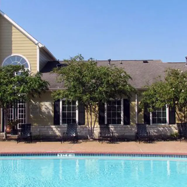 A clear blue swimming pool in front of a yellow building with a stone chimney, surrounded by greenery.