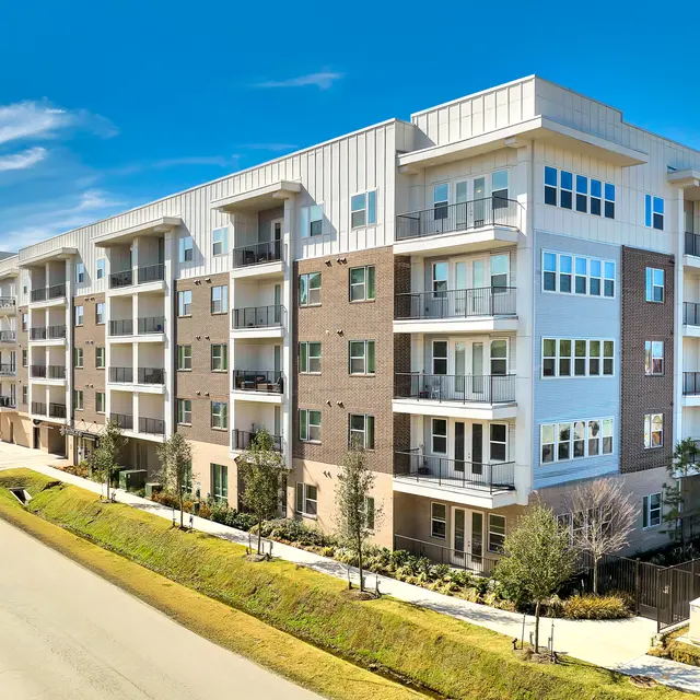 A modern apartment building with multiple floors, featuring a mix of brick and glass balconies. The building is set alongside a wide road and landscaped areas.