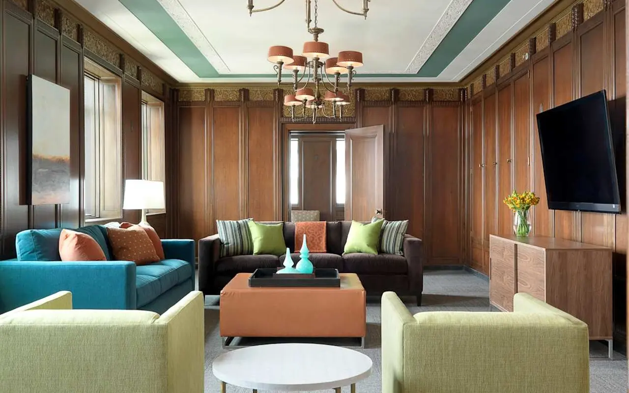 A stylish modern living room featuring a mix of colorful furniture, including blue, green, and orange seating arrangements, with wooden paneling on the walls. A chandelier hangs from the ceiling, and there is a flat-screen TV mounted on the wall.