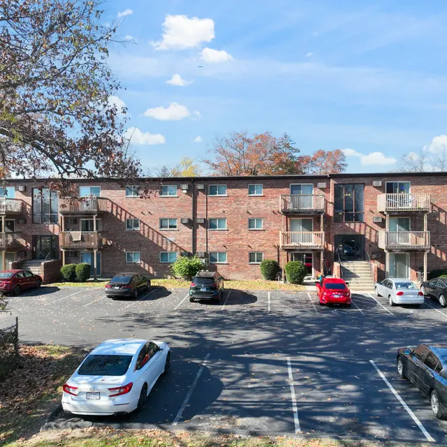 A multi-story brick apartment building with balconies, surrounded by trees in a parking lot with several cars parked nearby.