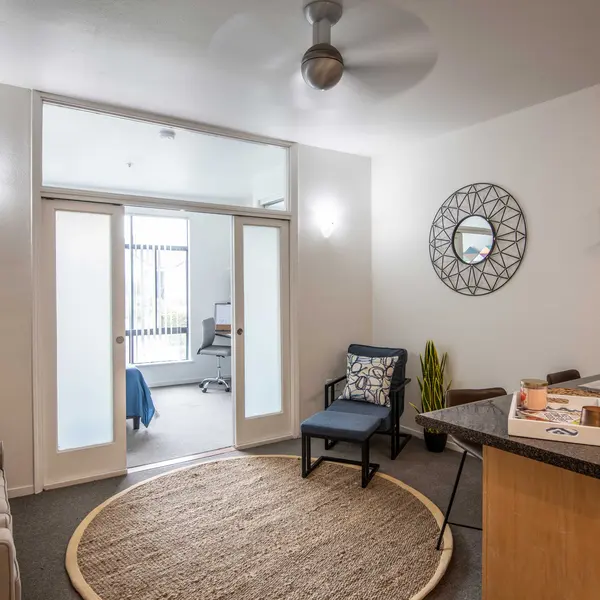 A cozy living area featuring a sofa with pillows, a round rug, a chair, and a dining area. Plants are placed around the room, and there is a large circular mirror on the wall.