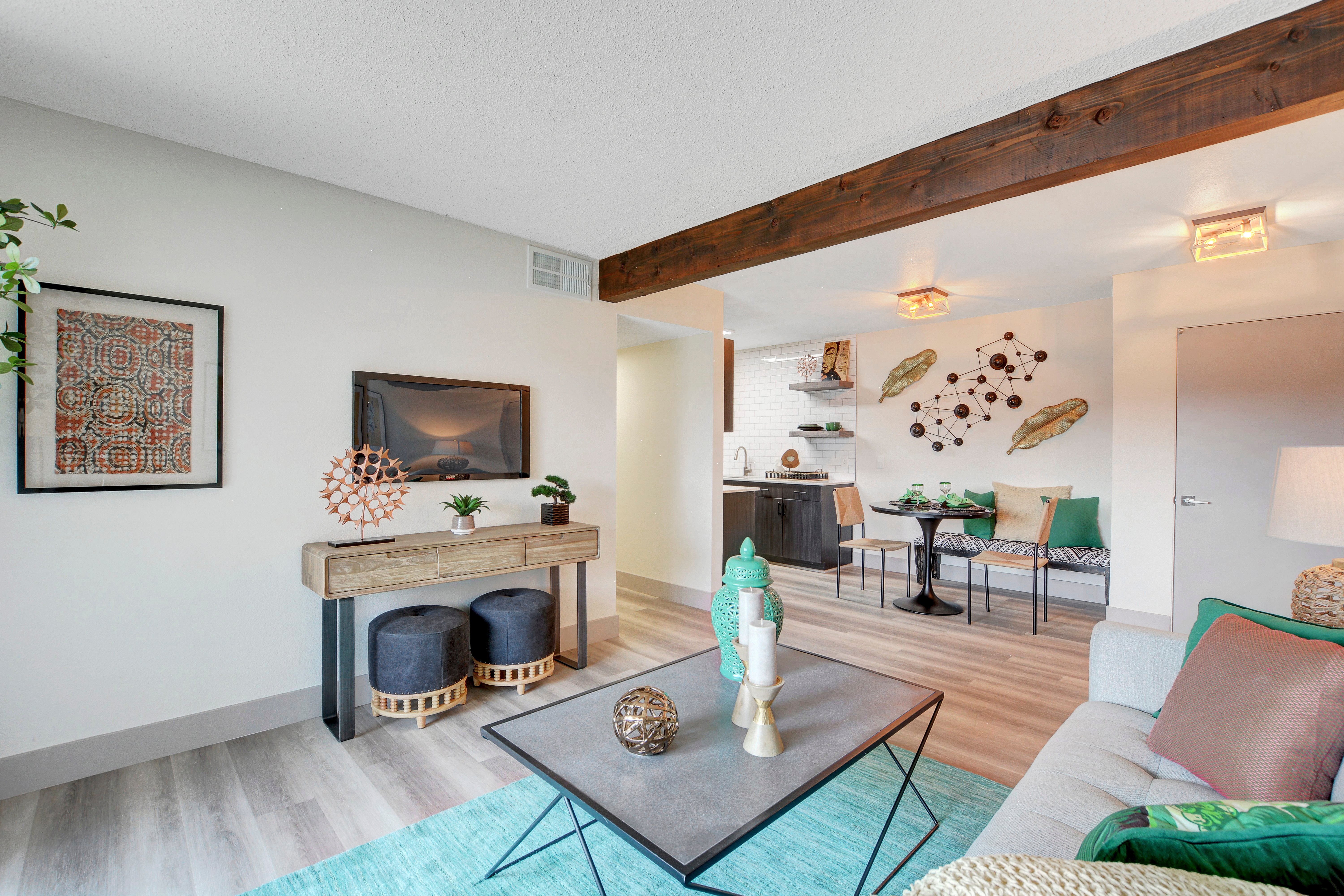 A stylish modern living room with light-colored walls, a wooden beam ceiling, and contemporary decor. It features a coffee table, a comfortable sofa with colorful cushions, and a decorative console table. Beyond, there's a dining area with a small table and chairs.