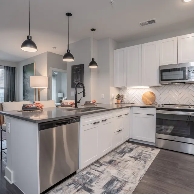 A modern kitchen featuring a large island, stainless steel appliances, and white cabinetry. The interior is well-lit with pendant lights and has a stylish decor with a mix of textures and colors.