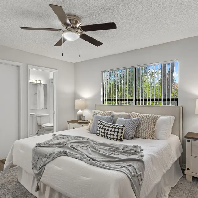 A modern bedroom featuring a double bed with cozy bedding, bedside tables with lamps, a ceiling fan, and a window revealing greenery outside.