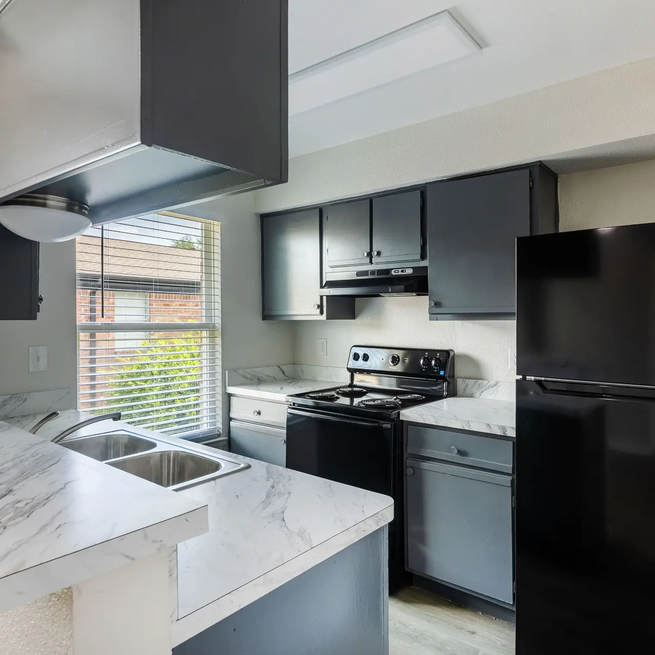 A modern kitchen featuring gray cabinets, a black refrigerator, and a black stove. The countertop is marble-like, and there is a window with blinds allowing natural light in.