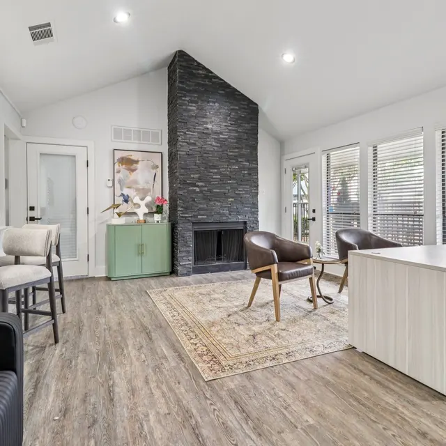 A modern living room featuring a black stone fireplace, large windows with blinds, a wooden floor, and a stylish rug. There is a desk with a computer and chairs placed around the room.