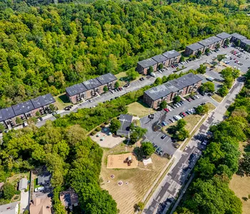 Aerial view of an apartment complex surrounded by trees. The complex features several buildings with solar panels on the roofs, a parking area, and a playground area.