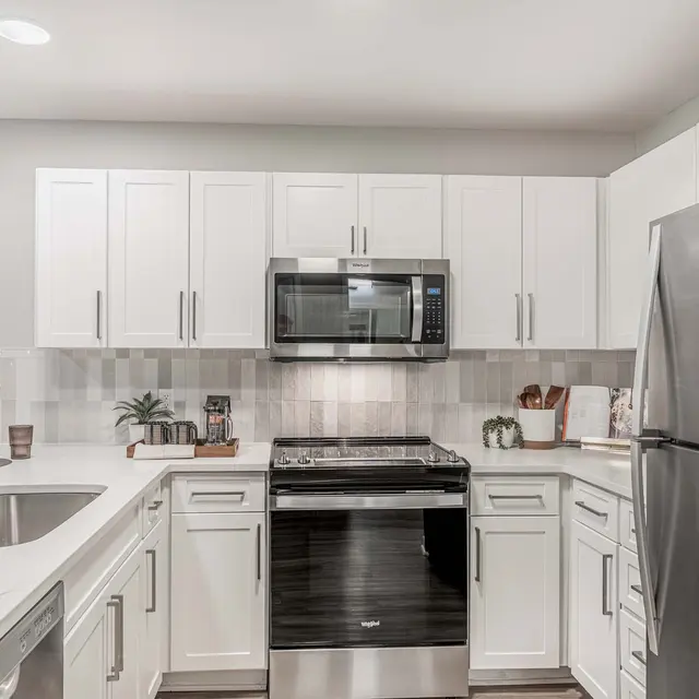 A modern kitchen featuring white cabinets, a stainless steel refrigerator, and a sleek microwave above a stove. The countertop is white with a large sink and a small plant on display.