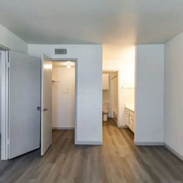Interior view of an apartment, featuring an open layout, light-colored walls, and wooden flooring. There's a doorway leading to what appears to be a bathroom and a small area that may serve as a kitchen in the background.