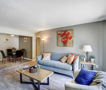 A modern living room featuring a blue sofa, coffee table, and lamp, with a dining area visible in the background.