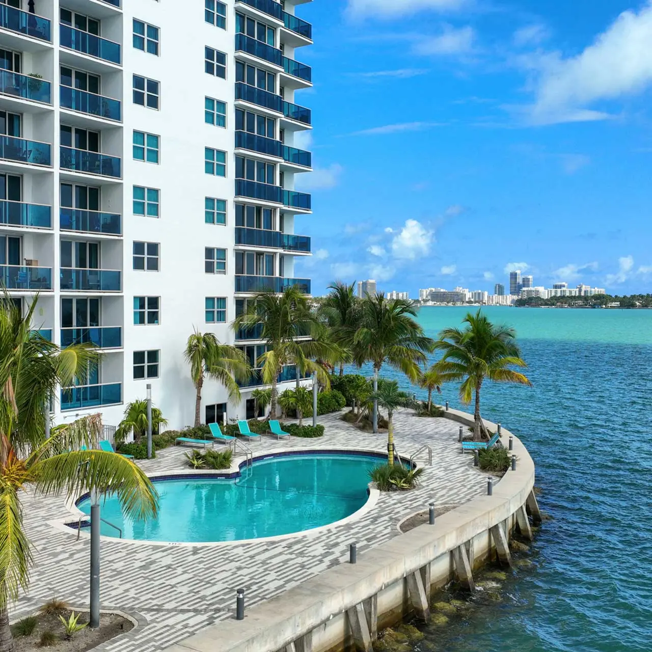 A waterfront condominium building with a swimming pool and palm trees, overlooking the water.