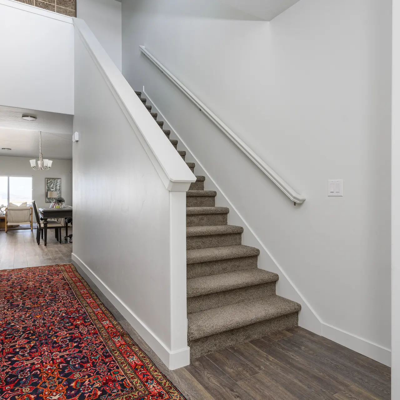 A well-lit interior staircase and hallway with a carpet runner. The stairs have a white railing and are flanked by a light grey wall. There's a large open space visible at the end of the hallway, suggesting more rooms beyond.