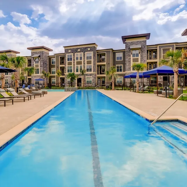 A clean swimming pool with lounge chairs alongside, surrounded by modern apartment buildings and palm trees under a partly cloudy sky.