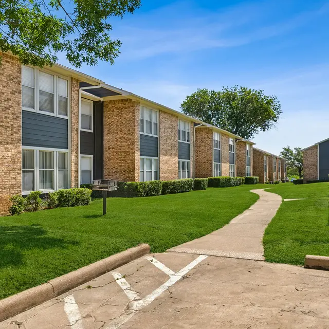 A view of a landscaped apartment complex with two buildings, green grass, and a winding pathway under a clear blue sky.