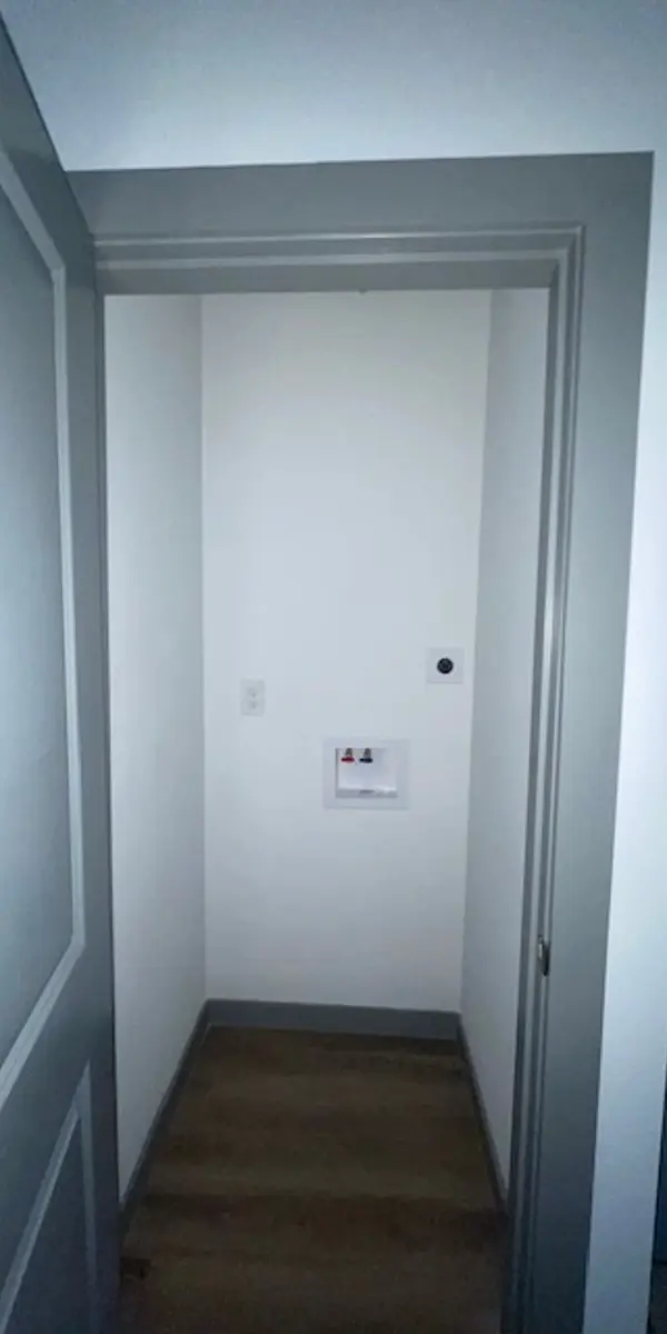 An empty, well-lit hallway with an open door at one end, featuring light-colored walls and wooden flooring.