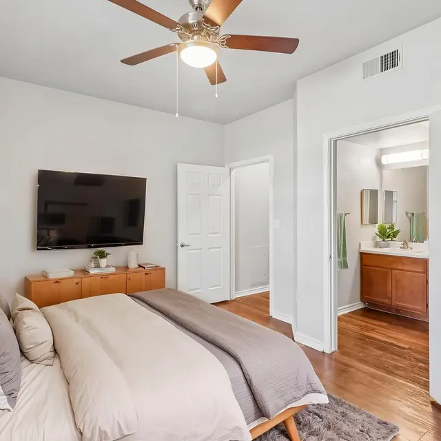 Bedroom (Staged) - Costa Almadena A stylish bedroom featuring a large bed, a TV mounted on the wall, a wooden dresser, and an attached bathroom. A ceiling fan is visible above, and a potted plant adds a touch of greenery.