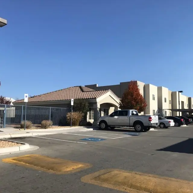 A view of a parking lot in front of a building with a fence. There are trees with red leaves suggesting autumn, and several parked cars.