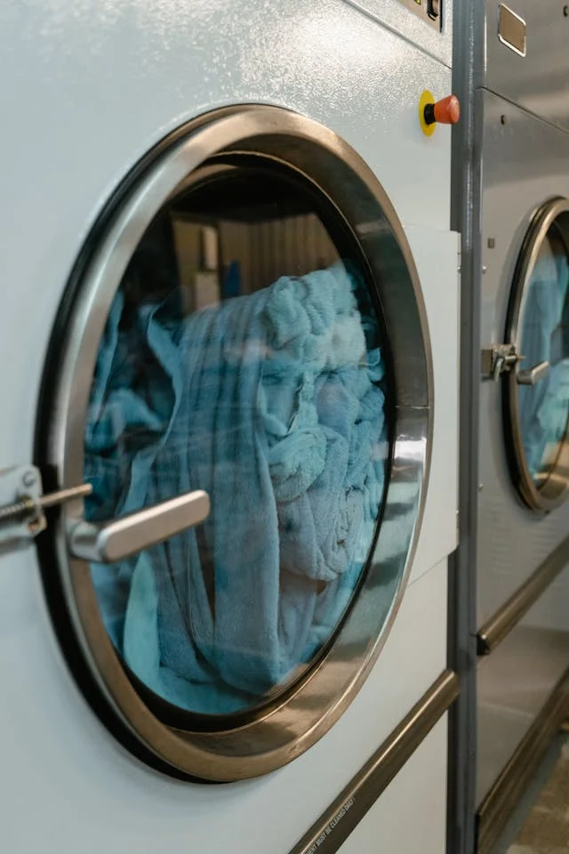 Blue Linens in a Commercial Washing Machine A close-up view of a washing machine's glass door showing blue bed linens inside. The machine is part of a row of commercial laundry machines.