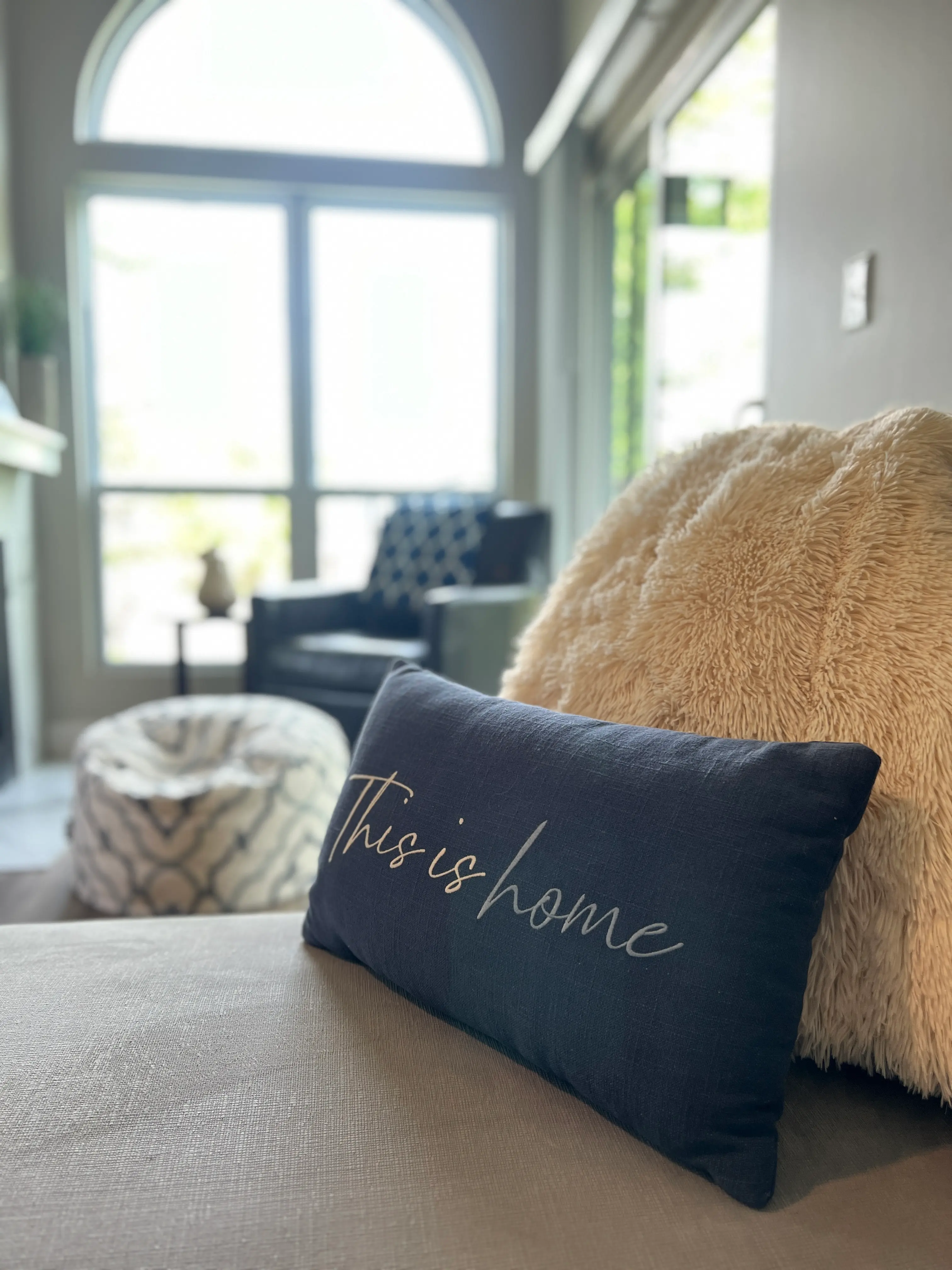A cozy living room with decorative pillows. A navy pillow with the text 'This is home' is foregrounded. There is a chunky white throw pillow and a patterned ottoman in view, along with a comfortable armchair and window allowing natural light to fill the space.