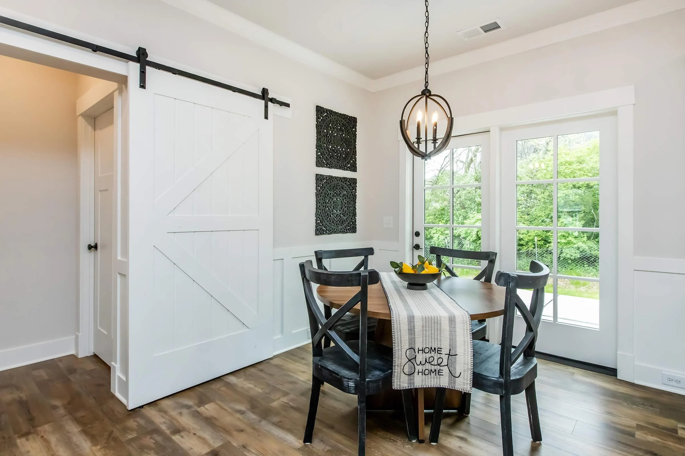 Modern Dining Room with Farmhouse Elements A modern dining area with a round wooden table, black chairs, and a stylish light fixture. There is a decorative table runner with the words 'Home Sweet Home' on it. A sliding barn door is partially visible, as well as large windows that look out onto greenery.