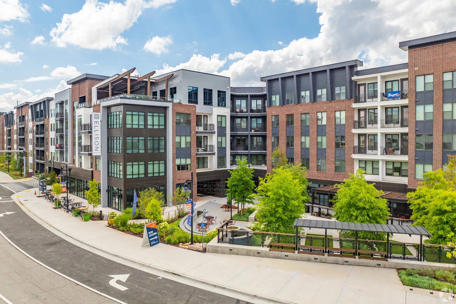 A modern apartment complex featuring a mix of brick and glass architecture, lush greenery and a well-maintained walkway.