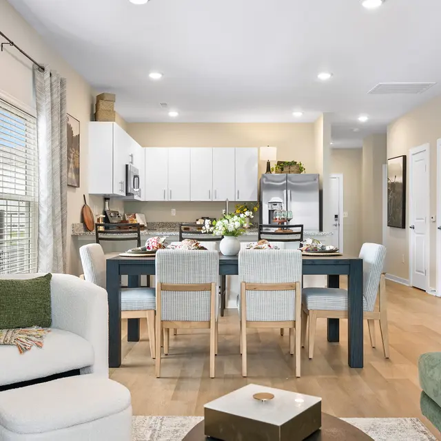 A bright, modern living space featuring a dining table set for a meal, surrounded by contemporary chairs. The kitchen area is visible in the background with white cabinetry and stainless steel appliances. Cozy seating arrangements include a green couch and a textured chair with a small table in the center. Natural light floods the space through large windows, enhancing the warm, inviting atmosphere.