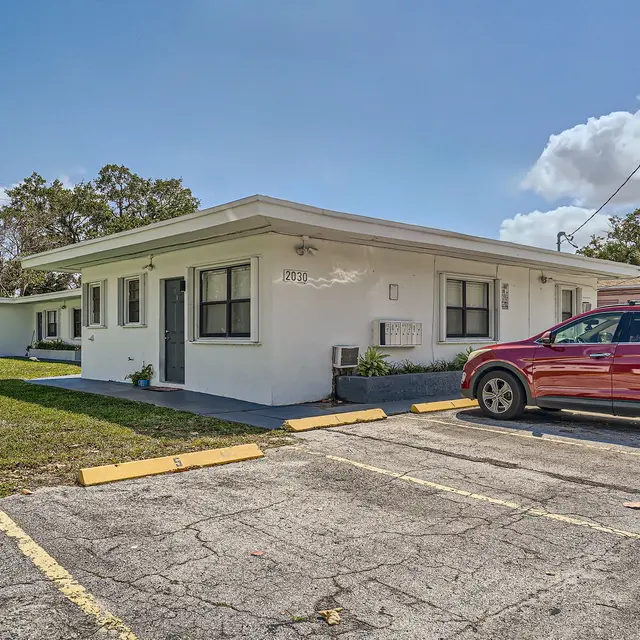 A view of a single-story apartment building with a red car parked in the foreground and grassy area nearby.