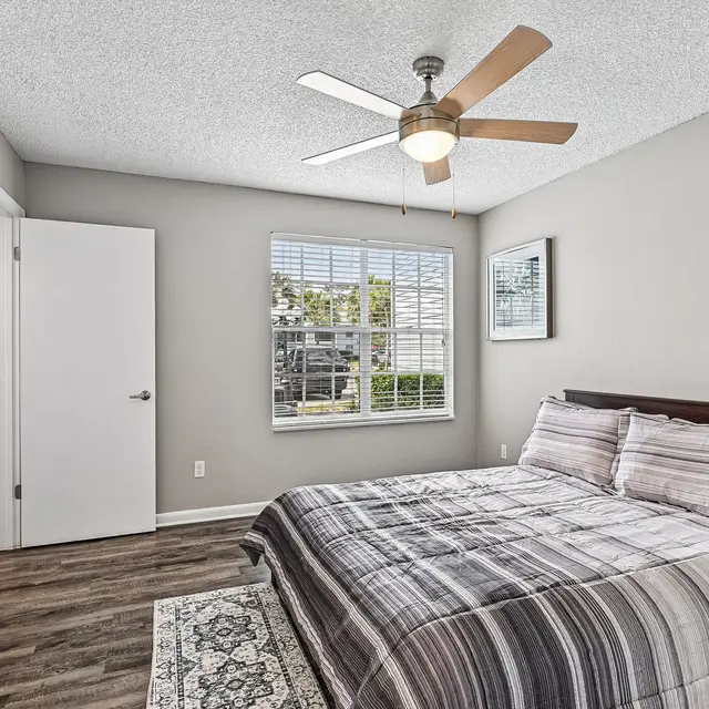 Contemporary Bedroom Design A modern bedroom featuring a double bed with striped bedding, a ceiling fan, a large window with blinds, and light gray walls. There is a round mirror and a wooden door leading to another room.