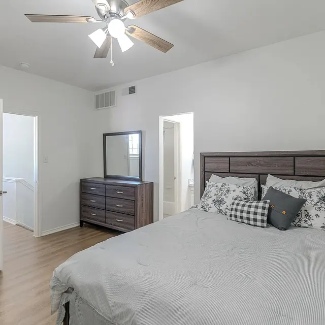 A well-lit bedroom featuring a bed with decorative pillows, a dark wood dresser, and a mirror. The flooring is laminate wood, and there are white walls. A door leads to a bathroom, showing a glimpse of the interior.