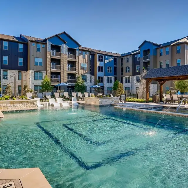 A luxurious apartment complex featuring a large swimming pool surrounded by lounge chairs and trees, with buildings in the background under a clear blue sky.
