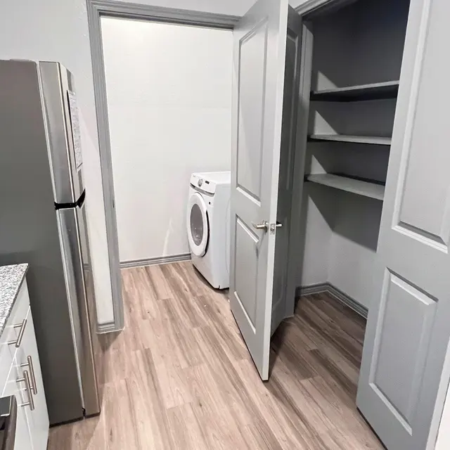 A small laundry area featuring a washer and space for shelving. The room is light-colored with wooden flooring and grey walls.