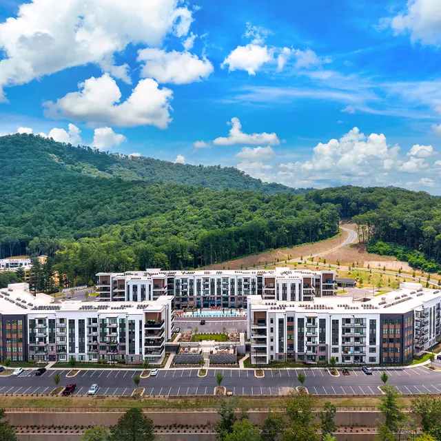 Aerial view of a modern apartment complex with multiple buildings surrounded by greenery and hills under a bright blue sky with clouds.