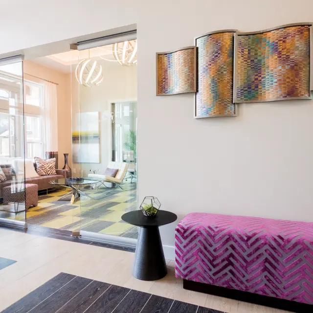 A modern lobby area showcasing a vibrant pink bench, a round black side table, and colorful wall art. A glass door leads to a bright living space beyond, with stylish furniture and large windows.