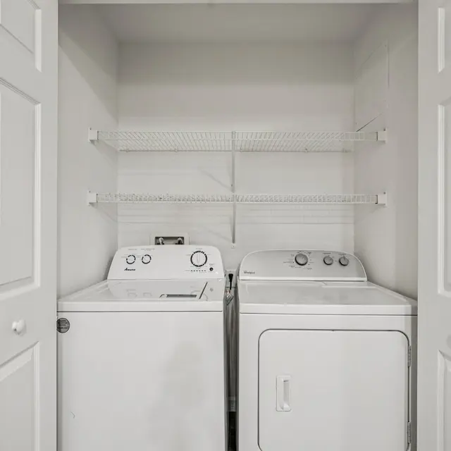 A compact laundry area featuring a washing machine and a dryer inside a closet with double doors. Above the appliances, there is a wire shelf for storage. The room is painted in light colors, giving it a clean look.