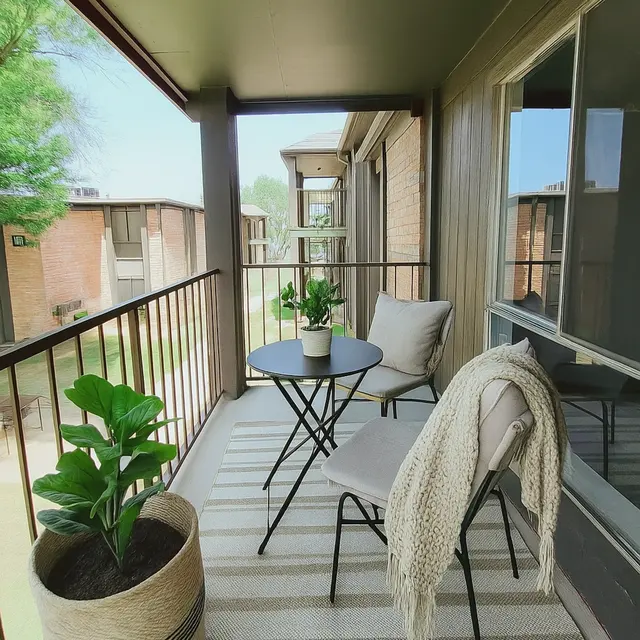A cozy balcony with a small round table and two chairs, accented by houseplants and a decorative throw. The balcony has a view of a green area and neighboring buildings.