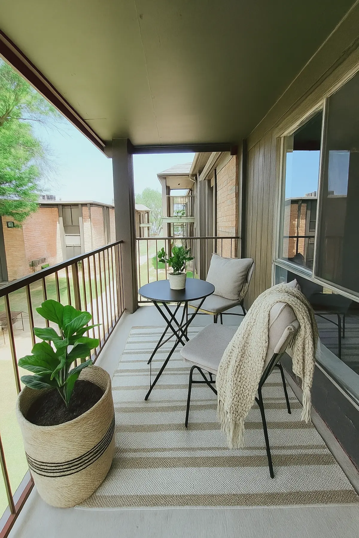 A cozy balcony with a small round table and two chairs, accented by houseplants and a decorative throw. The balcony has a view of a green area and neighboring buildings.