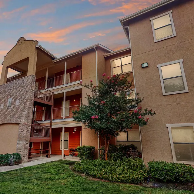 Residential apartment building with multiple levels, manicured lawns, and a colorful sunset in the background.
