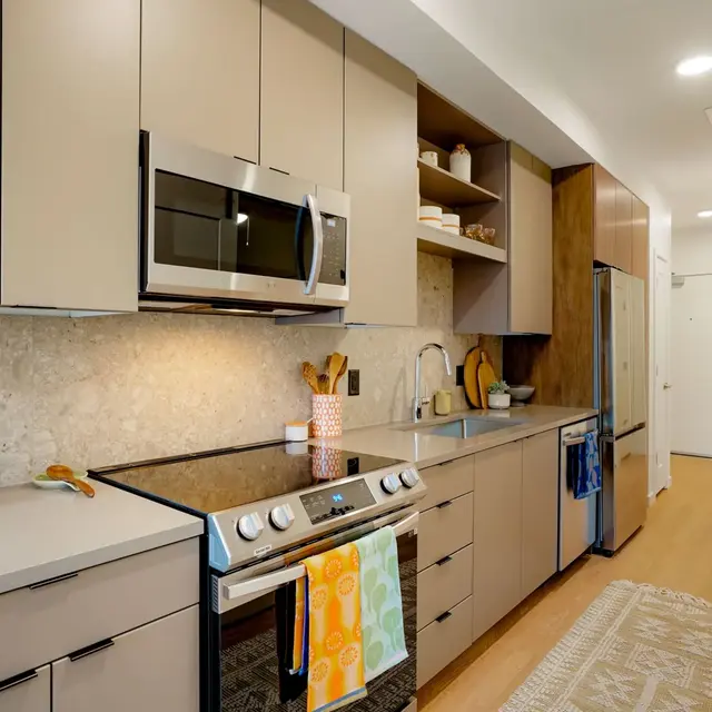 A modern kitchen with light-colored cabinets, stainless steel appliances, and a spacious layout. The countertop is sleek with decorative items placed on it, and a rug is laid on the floor.